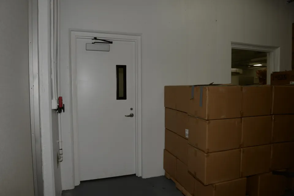Stacked brown cardboard boxes on a pallet beside a white door with a small window and metal handle in a warehouse corridor.