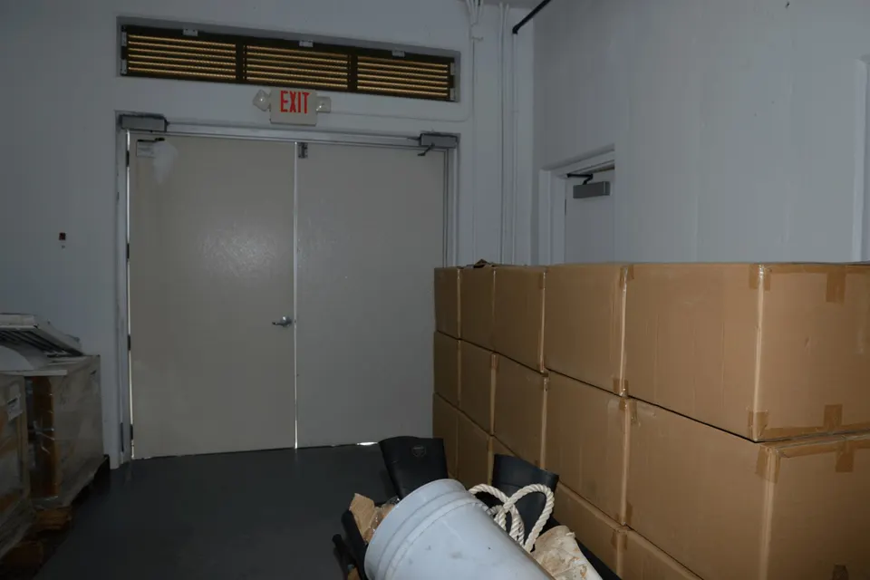 Stacked cardboard boxes fill the corner of a warehouse room beside white double doors with an illuminated exit sign above.