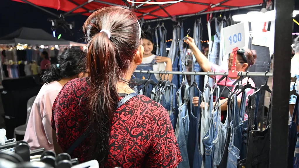 Shoppers browse hanging denim overalls and jeans at a red-roofed outdoor market stall, with a price tag reading 100 visible.