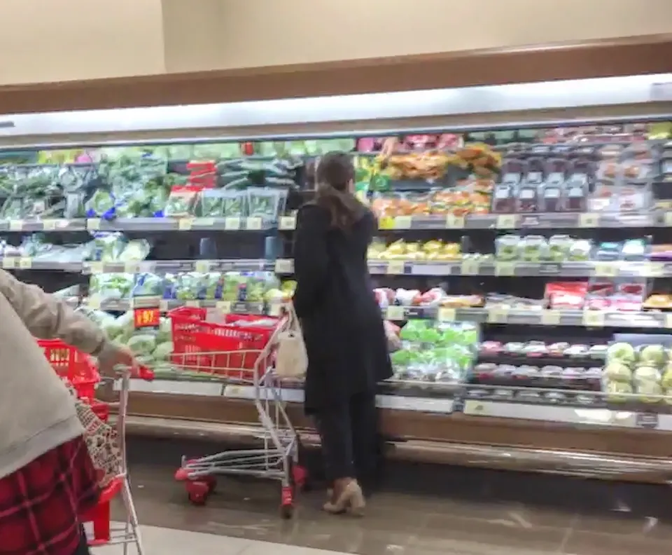 A woman in dark clothing looks at refrigerated produce shelves while a shopper with a red cart stands nearby in a supermarket.