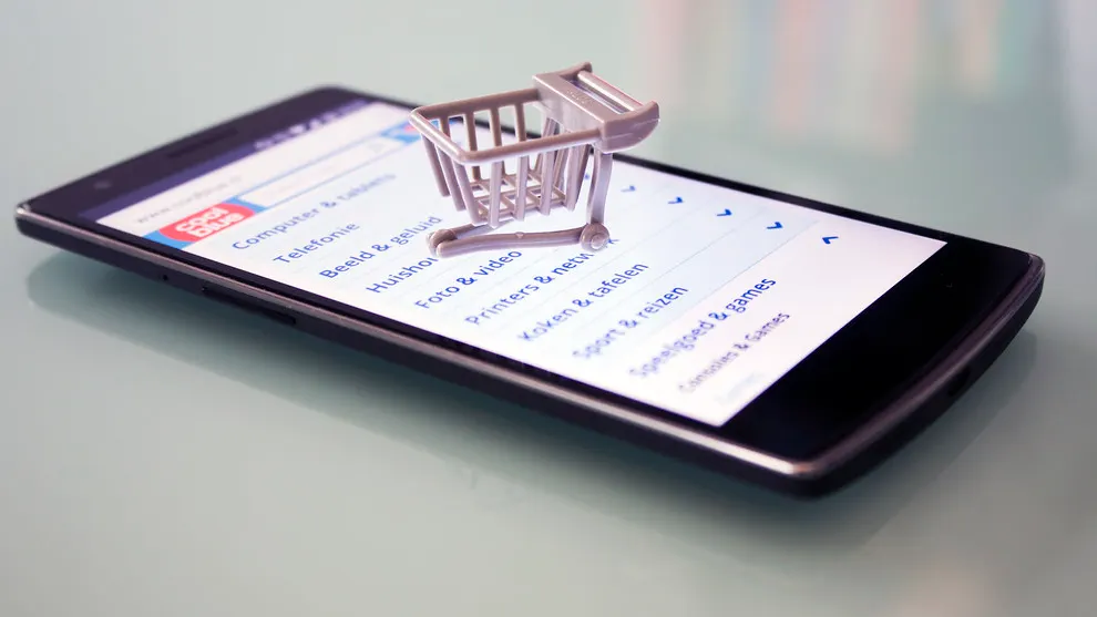 A smartphone on a glass table shows an online shop category menu, with a small toy shopping cart standing on the screen.