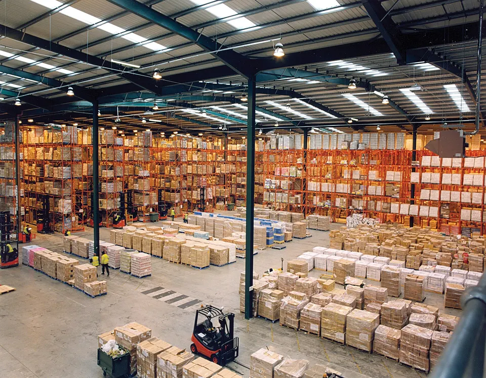 Large warehouse interior with tall orange pallet racks stocked with cardboard boxes, pallets on the floor, and a red forklift in the foreground.