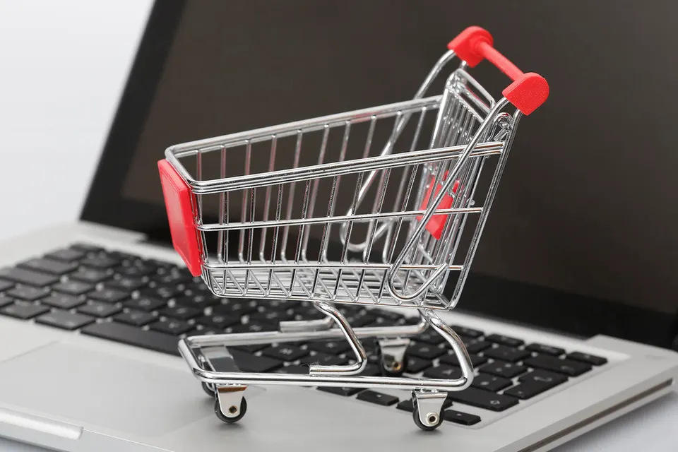 A miniature silver shopping cart with red handle and red seat sits on a silver laptop keyboard in front of a dark screen.