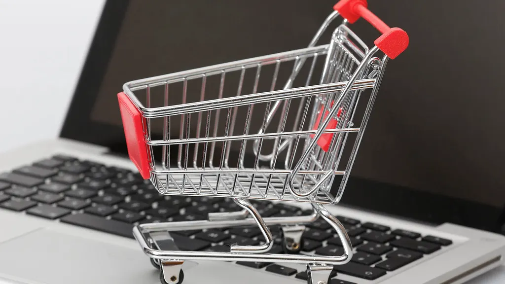 A miniature silver shopping cart with red handle and red seat sits on a silver laptop keyboard in front of a dark screen.
