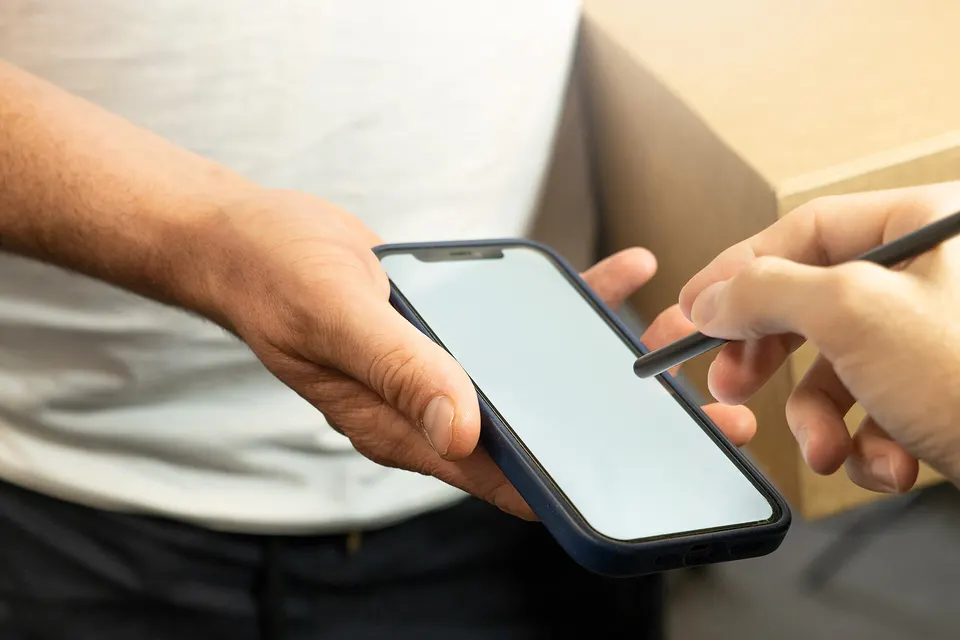 A man in a white t-shirt holds a dark blue smartphone while a hand holds a stylus over the screen, with a cardboard box in the background.