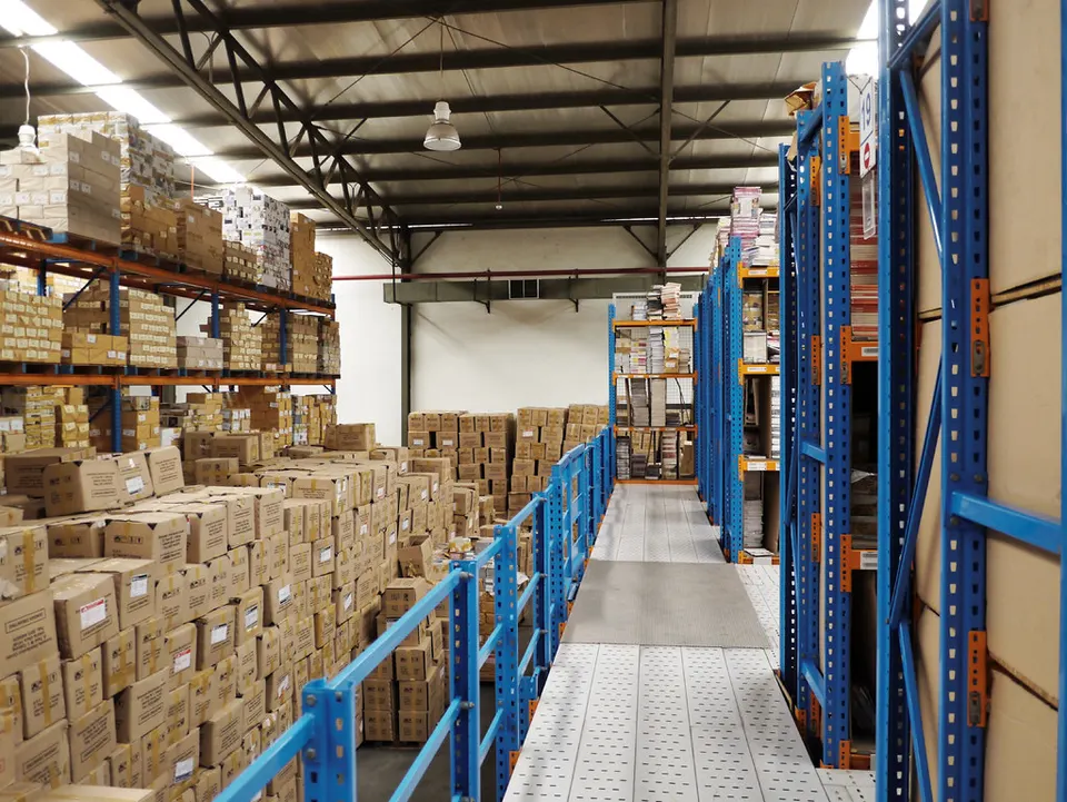 Warehouse aisle with blue metal pallet racking shelves stacked with cardboard boxes and packaged goods on both sides.